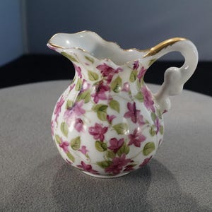 May include: A small white porcelain pitcher with a gold-trimmed handle and a floral pattern of pink and green flowers.