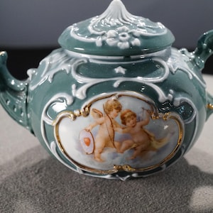 May include: A green and white porcelain teapot with a lid and a handle. The teapot features a decorative design of cherubs and flowers.