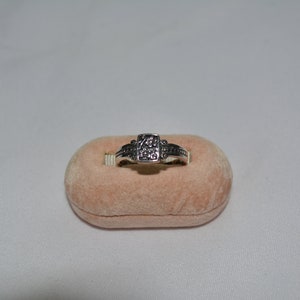 May include: A silver ring with a square-shaped stone set in the center. The ring is sitting on a light pink velvet cushion.