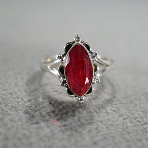 May include: A silver ring featuring a large, oval-cut, deep red gemstone. The gemstone is set in a decorative bezel with black accents and surrounded by a split shank band. The ring's design is ornate and eye-catching.