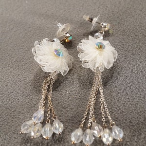 May include: A pair of dangle earrings with white flower-shaped charms and crystal accents. The earrings have multiple strands of gold chains with clear crystal beads.