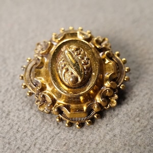 May include: A gold-toned oval brooch with a detailed floral design. The brooch has a raised center with a swirl pattern.