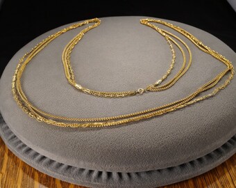 May include: A layered gold-tone necklace with three strands of varying chain styles. The chains are draped on a gray surface, showcasing the different textures and designs of the jewelry. The necklace is a classic accessory.