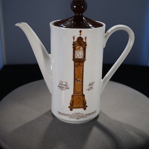 May include: White ceramic coffee pot with a brown lid and a brown image of a grandfather clock with the text "Hollandse staande klok" and "Anno 1750".