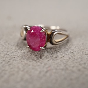 May include: A silver ring with a pink oval-shaped gemstone set in the center. The ring has a simple, elegant design with two open loops on either side of the stone.