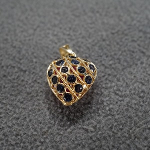May include: A gold-toned heart-shaped pendant with a filigree design. The pendant is encrusted with numerous dark blue gemstones. The background is a textured gray surface.