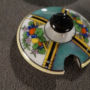 May include: A vintage porcelain lid with a teal, yellow and black geometric design. The lid features a floral motif with orange, yellow and purple flowers. The lid has a small spout for pouring.