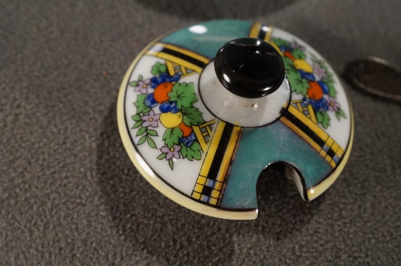 May include: A vintage porcelain lid with a teal, yellow and black geometric design. The lid features a floral motif with orange, yellow and purple flowers. The lid has a small spout for pouring.