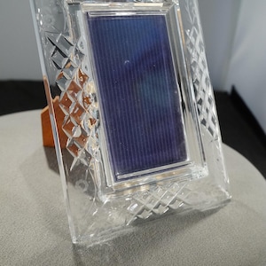 May include: A clear crystal picture frame with a rectangular photo area. The frame has a diamond-cut pattern and floral accents. The photo area has a blue and purple striped background. The frame is propped up on a wooden stand.