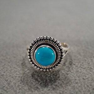 May include: A silver ring with a round, bright turquoise stone. The stone is surrounded by a detailed, beaded silver setting. The ring has a simple band and is set against a gray background.
