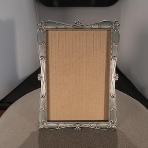 May include: A rectangular picture frame with a metallic silver finish and an ornate, scrolling border design. The frame features a beaded inner edge and a plain, light brown cardboard backing visible through the center opening.
