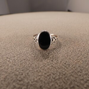 May include: A silver ring with an oval, black stone. The ring has a decorative openwork design on the sides of the band. The ring is set against a neutral background.