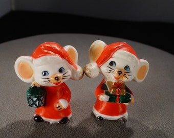 May include: Two white ceramic mice wearing red Santa hats and holding gifts. The mouse on the left is holding a green lantern, while the mouse on the right is holding a green box with a gold bow.