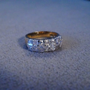 May include: A gold-toned ring with a band of rectangular and round-cut clear stones. The ring has a double-row design of sparkling stones, creating a dazzling effect. The ring is set against a gray background.