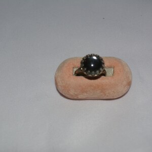 May include: A black gemstone ring with a gold setting. The ring is displayed on a pink velvet cushion.