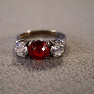 May include: A silver ring with a large red gemstone and two smaller clear gemstones.
