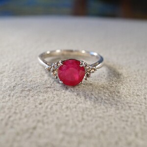 May include: A silver ring featuring a large, round, vibrant red gemstone as the centerpiece. The ring is accented with smaller, clear gemstones on either side of the main stone, set against a light-colored background.