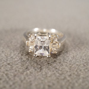 May include: A silver ring with a large, clear, square-shaped gemstone surrounded by smaller, clear gemstones.