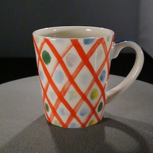 May include: A white ceramic mug with a red, blue, green, and yellow geometric pattern. The mug has a handle and is sitting on a gray surface.