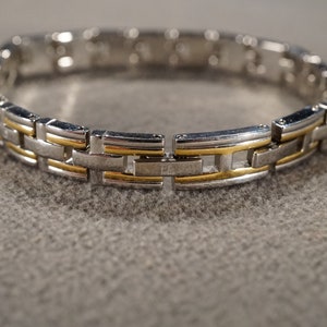 May include: A silver and gold tone bracelet with a link chain design. The bracelet is made of metal and has a polished finish.
