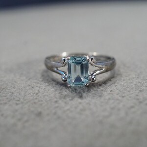 May include: A silver ring with a light blue gemstone set in an emerald cut. The ring has a delicate design with two curved prongs on either side of the stone.