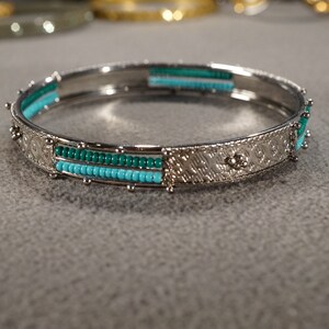 May include: A silver bangle bracelet with turquoise and green beads. The bracelet has a decorative design on the metal.
