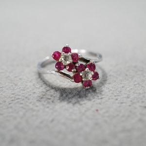May include: A silver ring with two flower-shaped settings, each with a cluster of red and white gemstones.