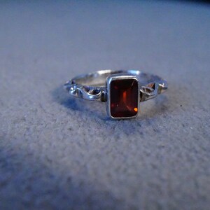 May include: A silver ring with a red gemstone set in a rectangular shape. The ring has a delicate, intricate design.