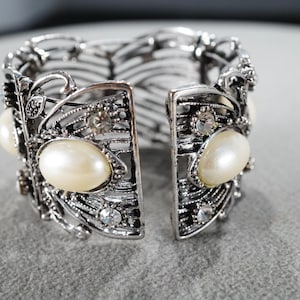 May include: A silver bracelet with a delicate, intricate design featuring faux pearls and rhinestones. The bracelet is open on one side and has a hinged closure.
