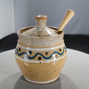 May include: A ceramic sugar bowl with a lid and a small wooden spoon. The bowl is tan with a white band and a blue wave design. The lid has a small handle. The spoon is inserted into the bowl.