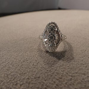 May include: A silver-toned ring featuring a marquise-shaped head with three prominent round stones arranged vertically in the center, surrounded by a halo of smaller pavé-set stones and additional baguette-cut stones radiating outward.