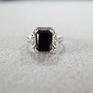 May include: A silver ring with a rectangular black gemstone. The ring has an ornate design around the gemstone and on the band. The ring is set against a light gray background.