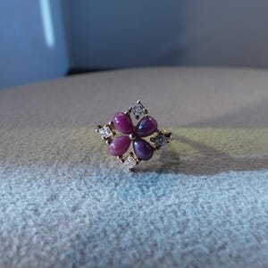 May include: A gold ring featuring a central floral cluster of four teardrop-shaped, deep pink cabochon stones arranged in a cross pattern, accented by four small, clear, square-cut stones set at the outer corners of the floral design.