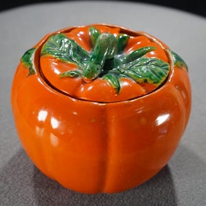 May include: Orange ceramic tomato-shaped jar with a lid. The lid is decorated with green leaves and a stem.