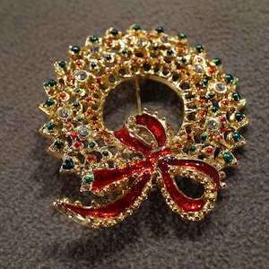 May include: A gold-tone Christmas wreath brooch with red enamel and green and clear rhinestones. The wreath has a red bow with gold trim.