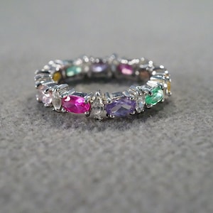 May include: A silver ring with a band of colorful oval-shaped gemstones. The stones are set in a row around the band and include pink, purple, green, and yellow.