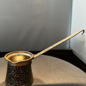 May include: A silver-colored, small coffee pot with an elongated handle. The pot features intricate, dark-colored etched designs around the body. The handle has a hook at the end. The pot is designed for brewing coffee.