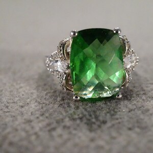 May include: A silver ring with a large, green, faceted gemstone surrounded by smaller, clear gemstones.