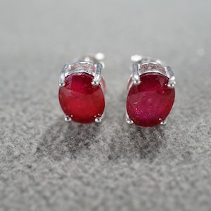 May include: A pair of oval-shaped ruby stud earrings. Each earring features a vibrant red gemstone set in a silver-colored metal. The earrings are displayed against a textured gray background.