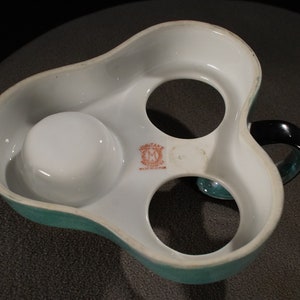 May include: A white ceramic egg holder with a teal green rim and a black handle. The holder has three compartments for eggs and a small compartment for salt or pepper. The holder has the words "MONTANE" and "MADE IN ENGLAND" printed on the bottom.