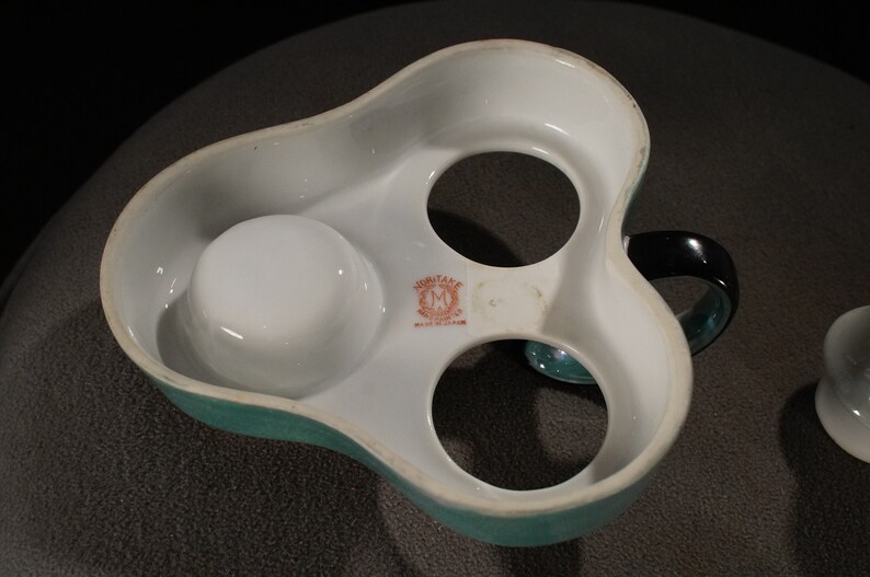 May include: A white ceramic egg holder with a teal green rim and a black handle. The holder has three compartments for eggs and a small compartment for salt or pepper. The holder has the words "MONTANE" and "MADE IN ENGLAND" printed on the bottom.