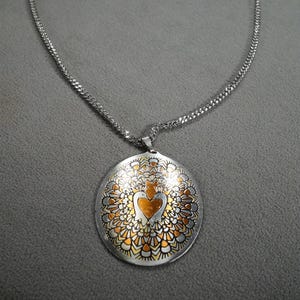 May include: A silver chain necklace with a large oval pendant. The pendant is made of silver and copper and features a heart design with two birds on either side. The pendant has a detailed mandala-like design around the heart.