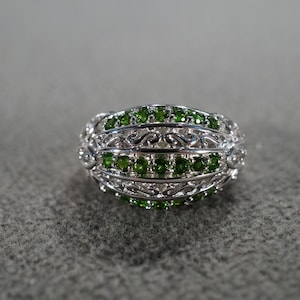 May include: A silver-toned ring with a detailed filigree design. The ring is adorned with rows of small, vibrant green gemstones. The gemstones are set within the intricate metalwork, creating a striking contrast.