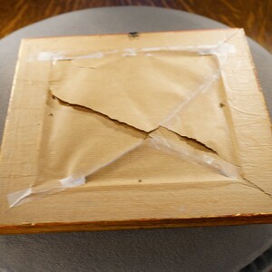 May include: The back of a wooden picture frame with a gold trim. The frame is cracked and has tape holding it together.