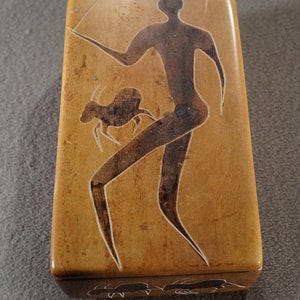 May include: A wooden box with a carved design of a person holding a spear and a small animal. The box is brown with a light brown background.