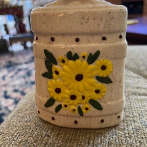May include: A white ceramic jar with a speckled finish and a floral design. The jar has a brown lid and is decorated with yellow flowers and green leaves.