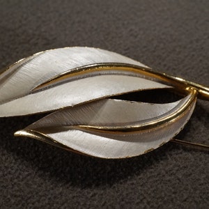 May include: A gold and white leaf-shaped brooch with a brushed metal finish.
