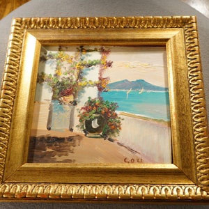 May include: A framed oil painting depicting a coastal scene with a white building, lush greenery, and a view of a blue ocean with sailboats and mountains in the distance. The painting is signed "C.O.U."