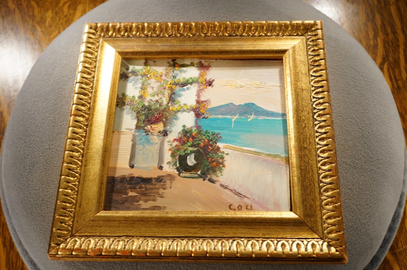 May include: A framed oil painting depicting a coastal scene with a white building, lush greenery, and a view of a blue ocean with sailboats and mountains in the distance. The painting is signed "C.O.U."