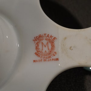 May include: Close-up of a white ceramic egg holder with a red and white Noritake logo that reads "Noritake Hand Painted Made in Japan".